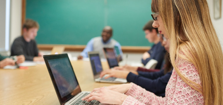 Students gather around a conference table typing on their laptops